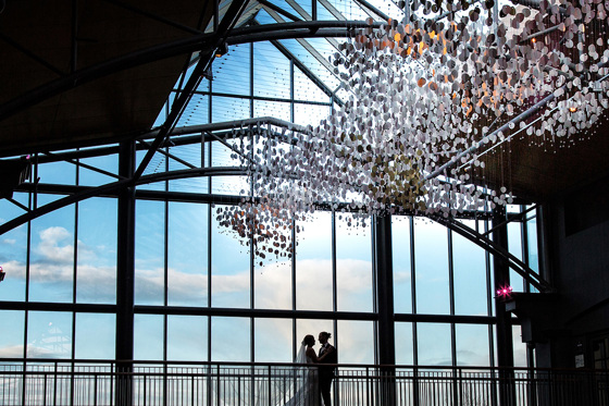 Bride and groom stand in front of large windows with beautiful sky and light fixture